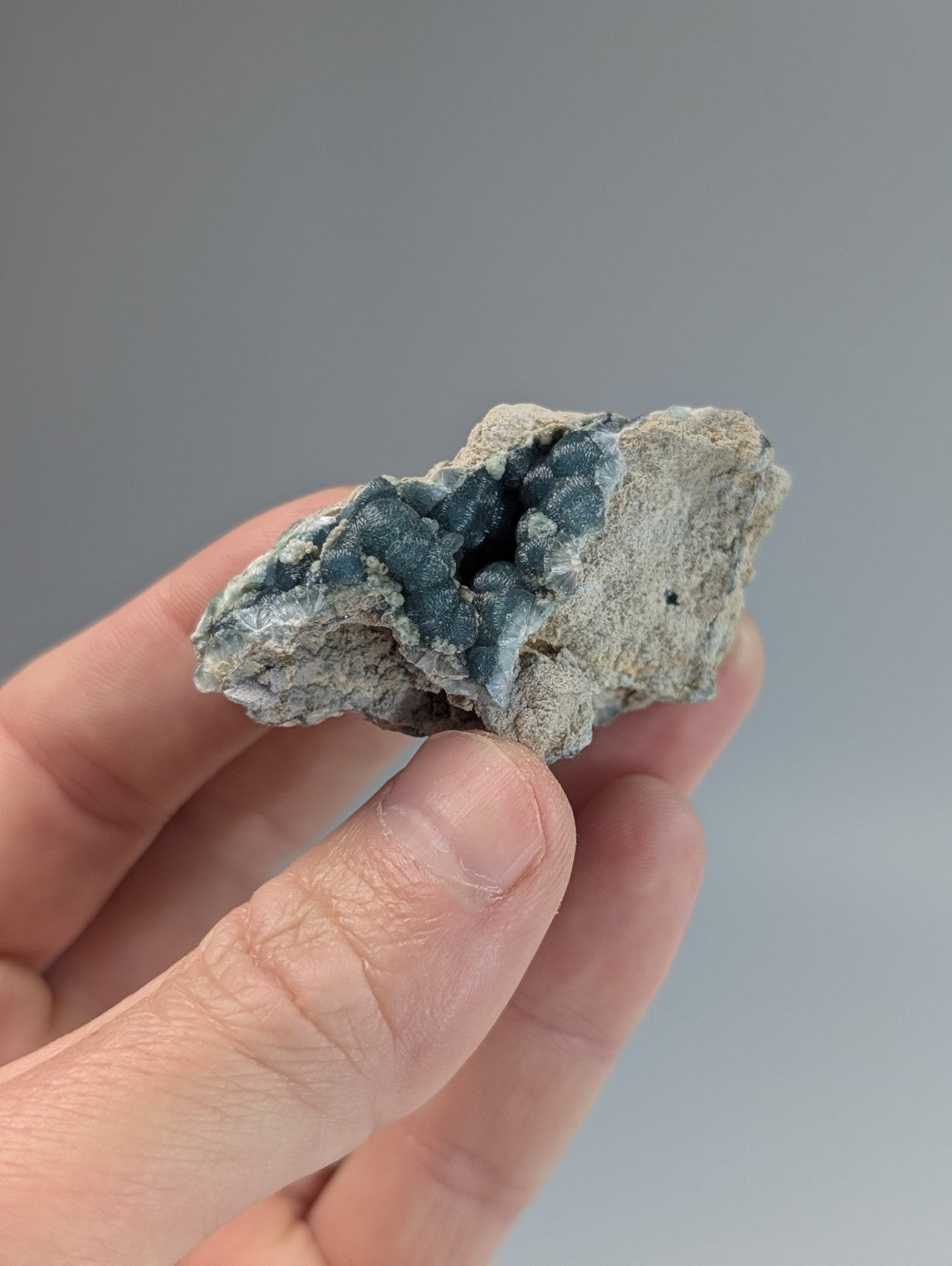 A hand holds a rough, irregularly shaped mineral specimen featuring a striking contrast between its pale gray matrix and the deep blue-green crystalline formations.