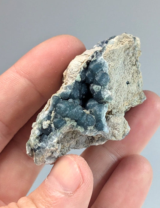 A hand holds a natural geode with a striking blue-green crystalline interior contrasting against its rough, grayish rock matrix.