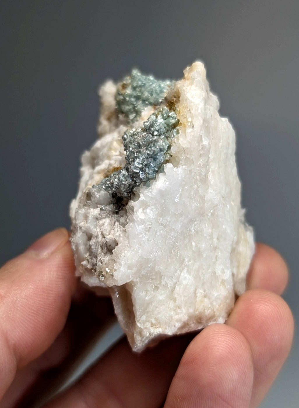 A hand holds a white quartz specimen with blue green cookeite and metallic blue green pyrite crystals from arkansas