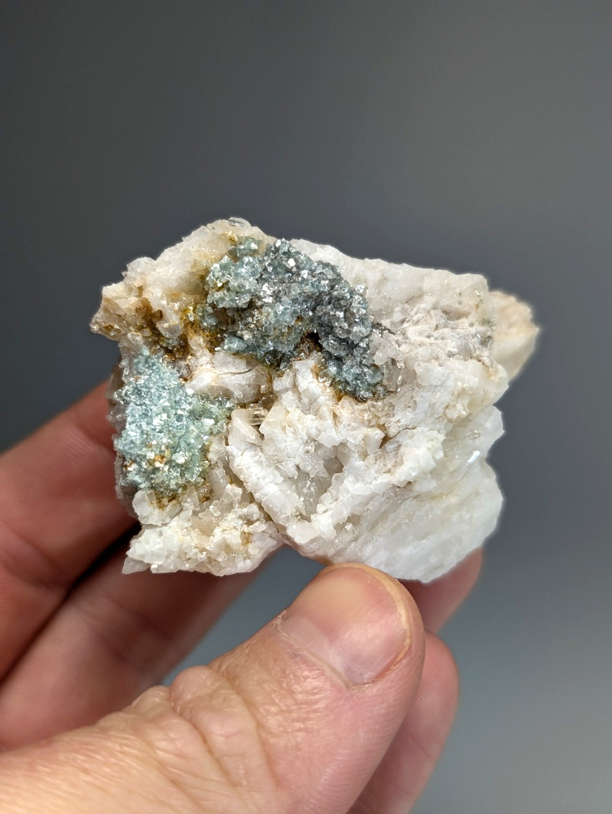 A hand holds blue green cookeite on white quartz with metallic gray and light blue crystals rare uv reactive mineral