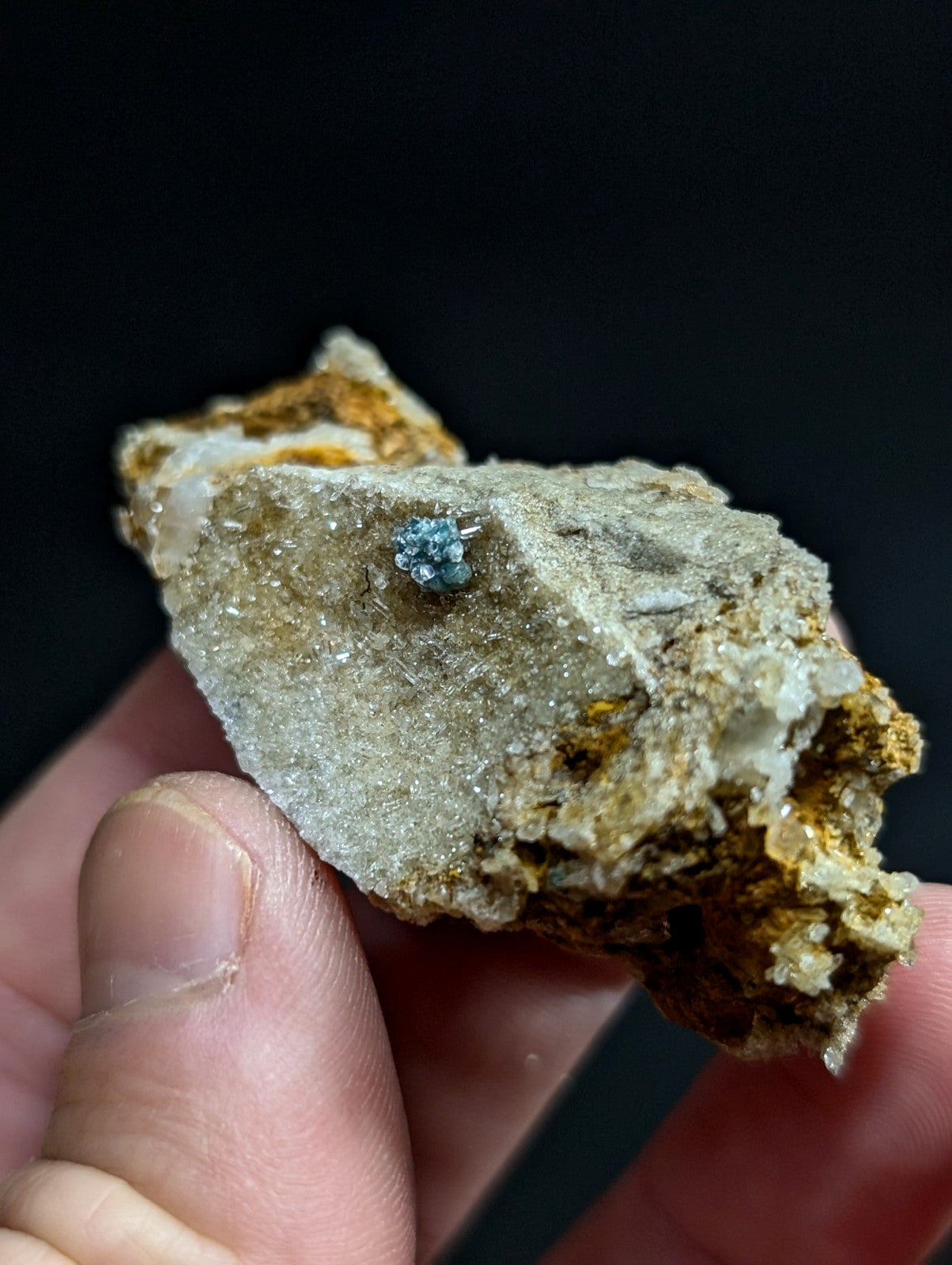A small, faceted blue gemstone nestled in a rough, textured rock matrix with patches of orange and white mineral veins.