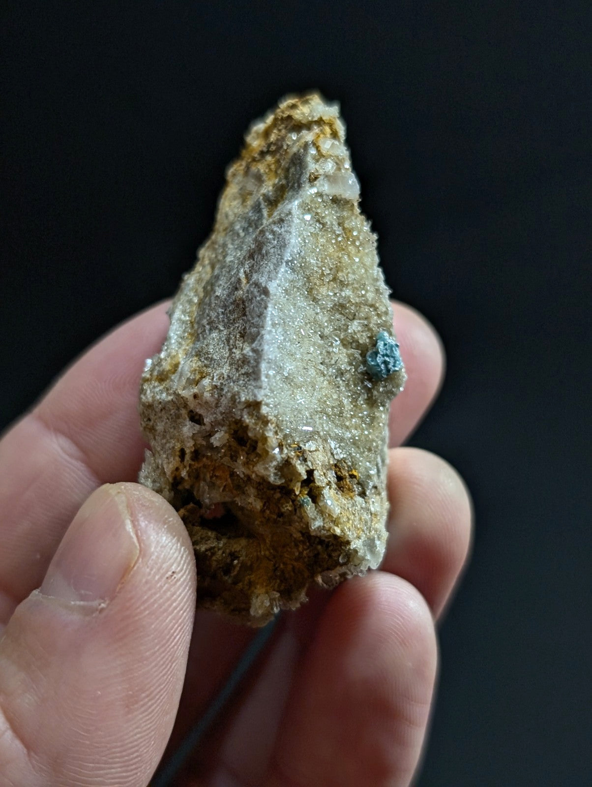 A rough, irregularly shaped mineral specimen with a mix of earthy brown and gray tones, featuring small crystalline formations and a distinct patch of vibrant blue-green on its surface.