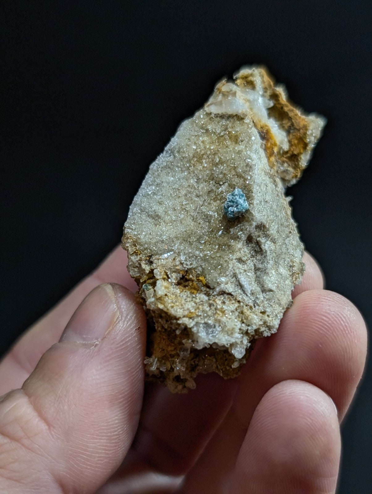 A hand holds a rough, irregularly shaped mineral specimen with a grayish-white base and patches of yellow-orange oxidation, featuring a small, distinct blue crystalline inclusion.