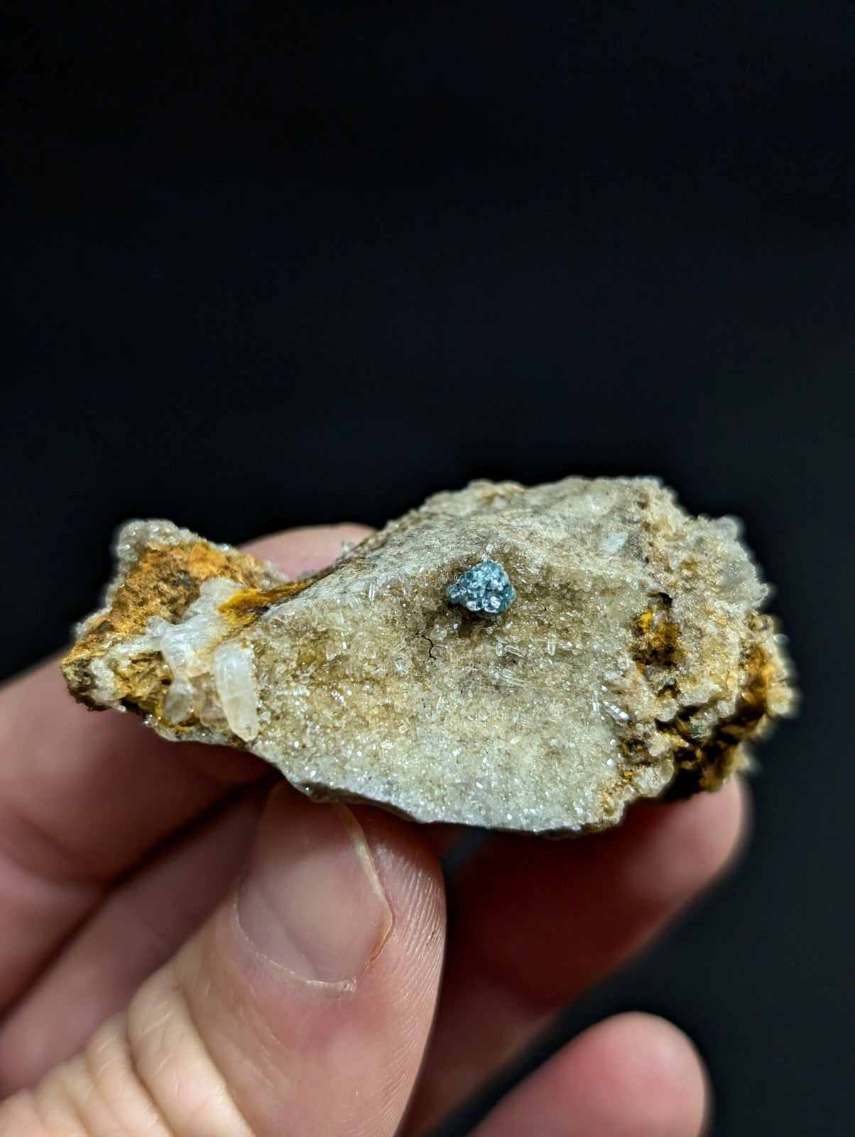 A hand holds a rough, irregularly shaped rock with a speckled gray and tan surface, featuring a small, dark blue-green mineral embedded in its center.