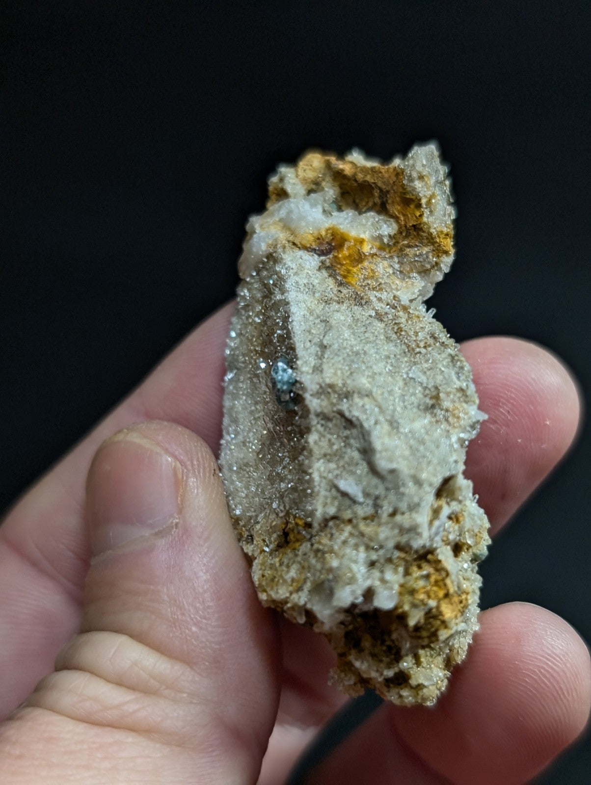 A hand holds a rough, irregularly shaped mineral specimen featuring translucent white crystals with patches of orange-yellow oxidation and a small embedded blue gemstone.
