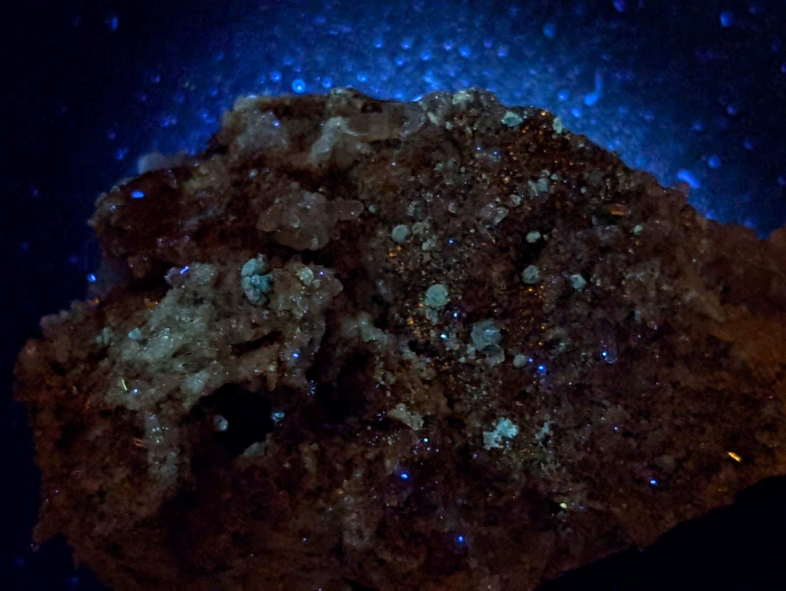 A rugged, earthy mineral specimen with a textured surface featuring translucent crystals and specks of glowing blue under ultraviolet light.