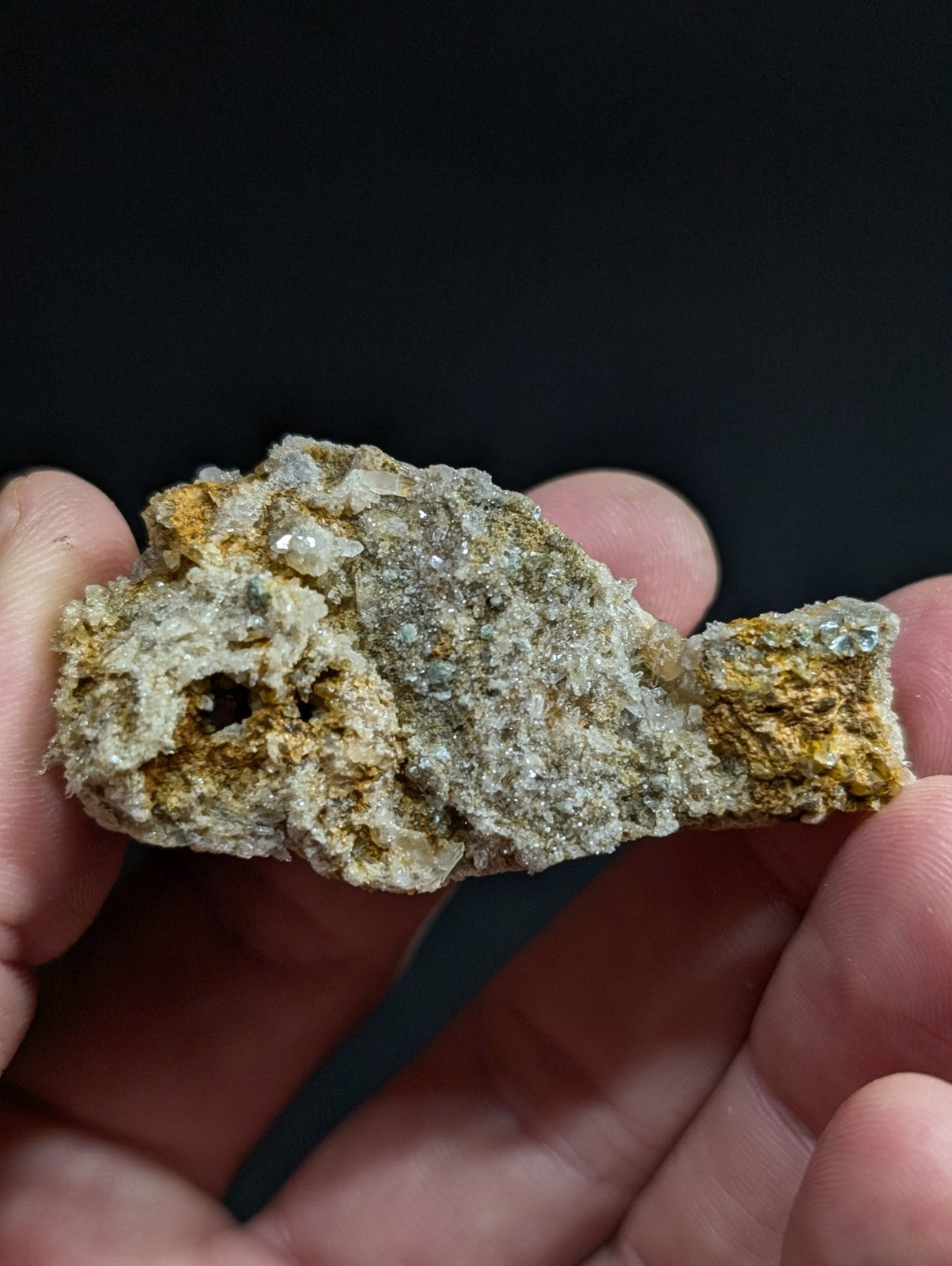 A hand holds a rough, irregularly shaped mineral specimen featuring patches of yellowish-orange and grayish-white crystalline formations.