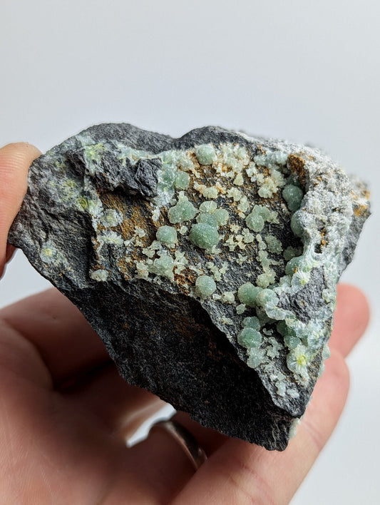 A hand holds a dark gray rock specimen featuring clusters of translucent, light green crystals embedded in its surface.