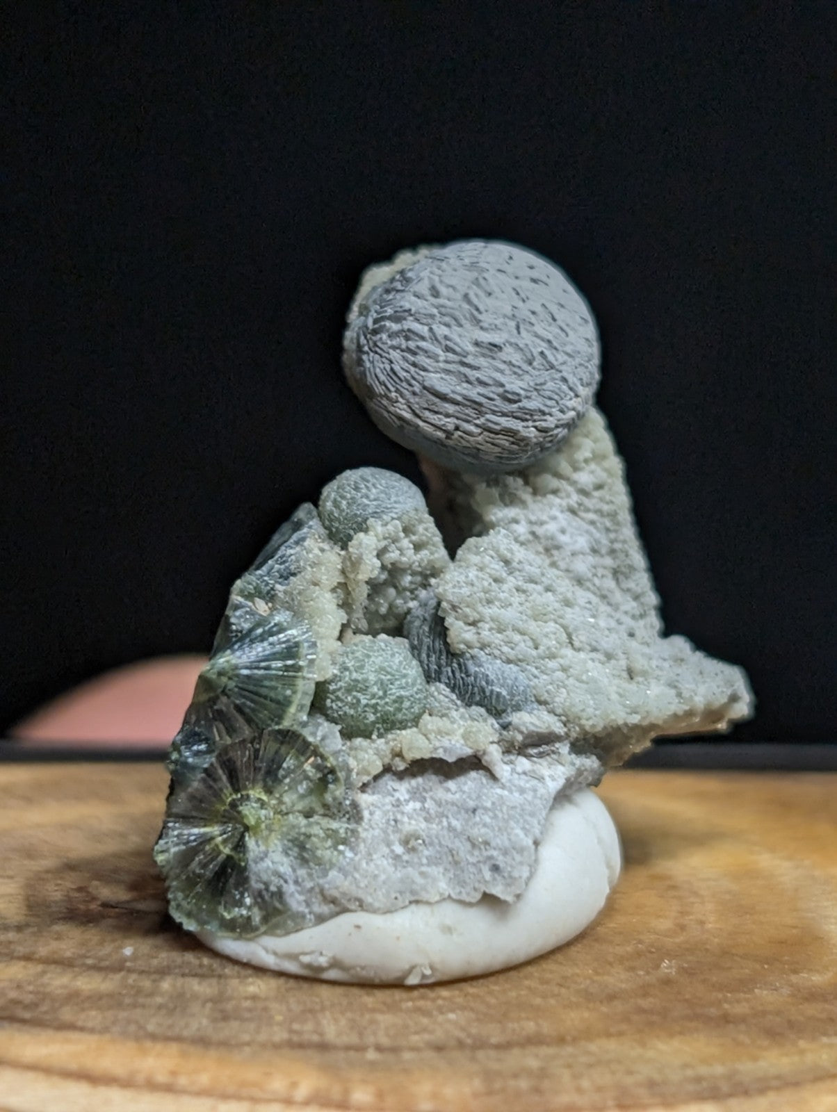 A gray, textured mineral specimen featuring a spherical formation atop layered crystalline structures with greenish hues and intricate patterns.