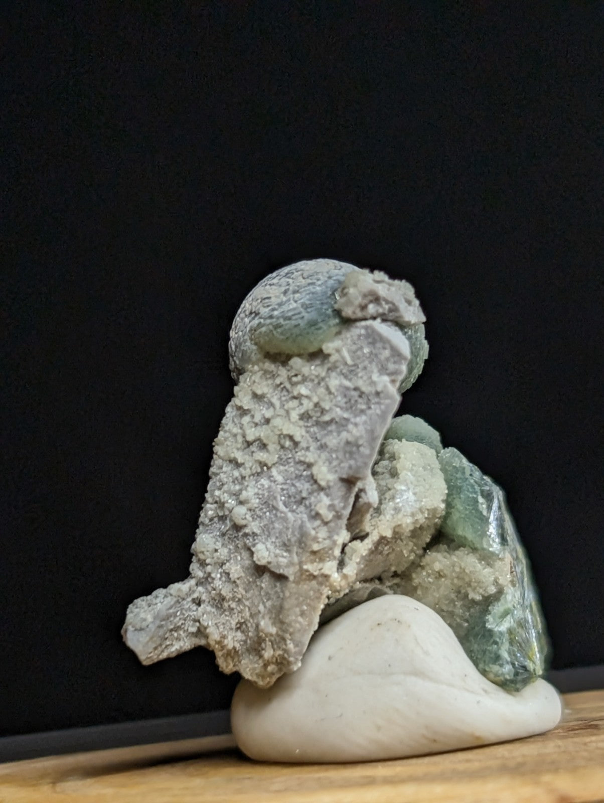 A textured, grayish-green mineral specimen with crystalline formations rests on a smooth white base.