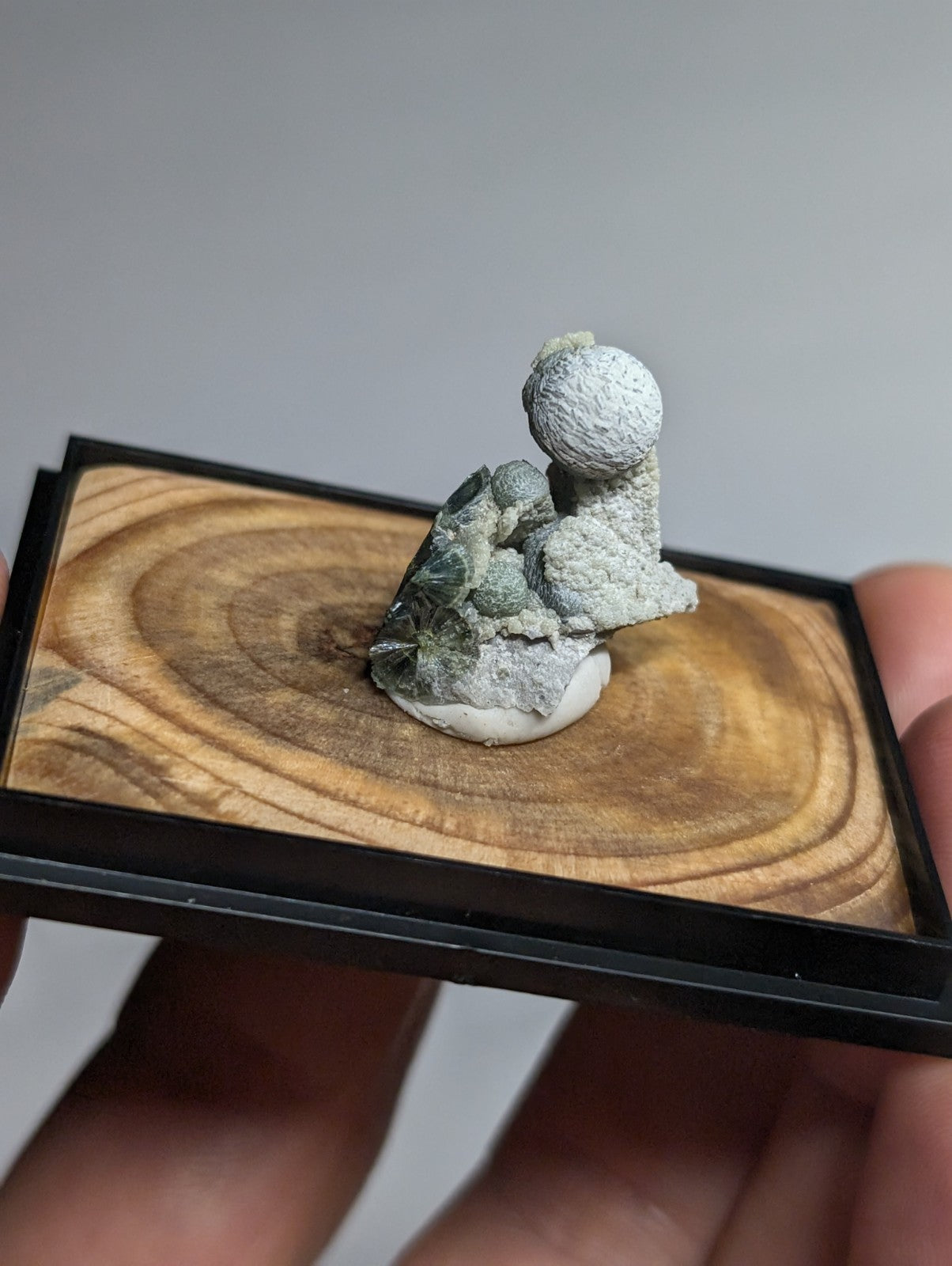 A miniature gray stone sculpture of a figure holding a sphere, displayed on a wooden base within a black frame.