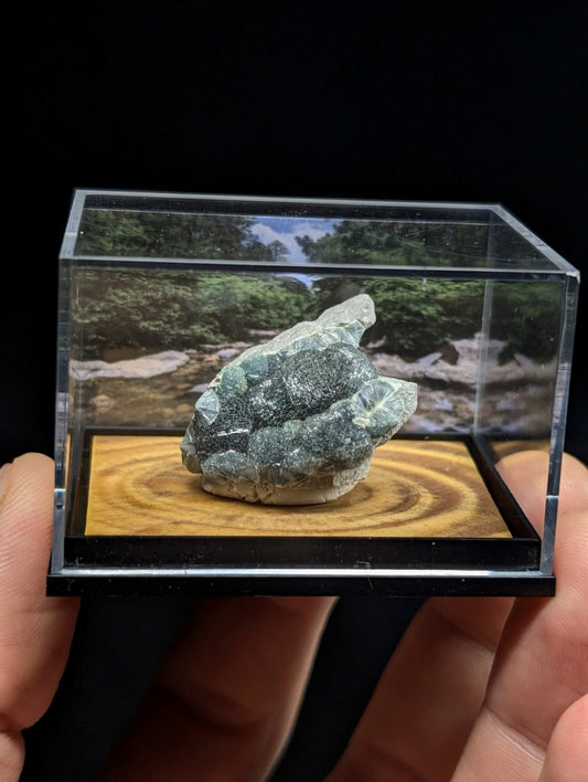 A textured, greenish-gray mineral specimen with a rough, irregular surface sits inside a clear acrylic display case.