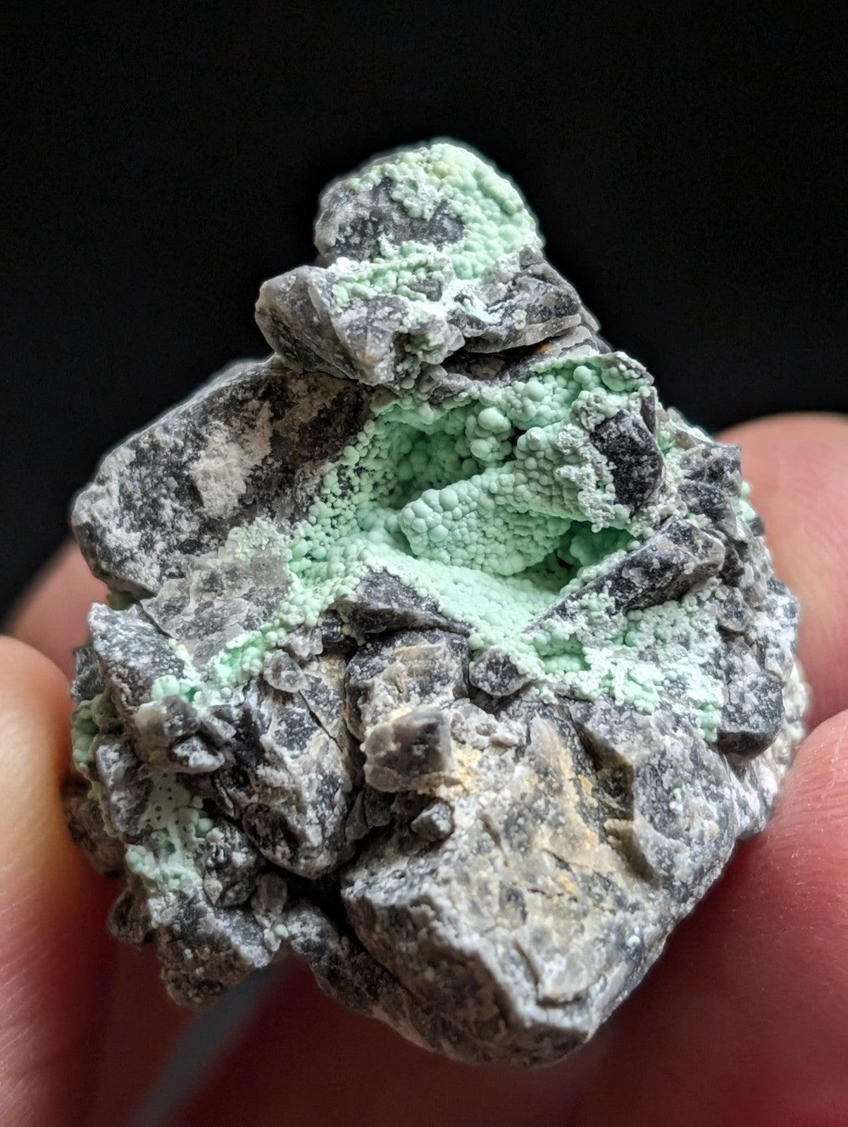 A hand holds a rough, irregularly shaped mineral specimen featuring dark gray rock with patches of vibrant light green crystalline growths.