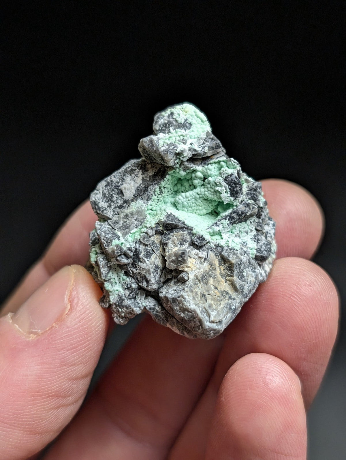 A hand holds a rough, irregularly shaped mineral specimen featuring patches of vibrant green patina over a dark gray rock base.