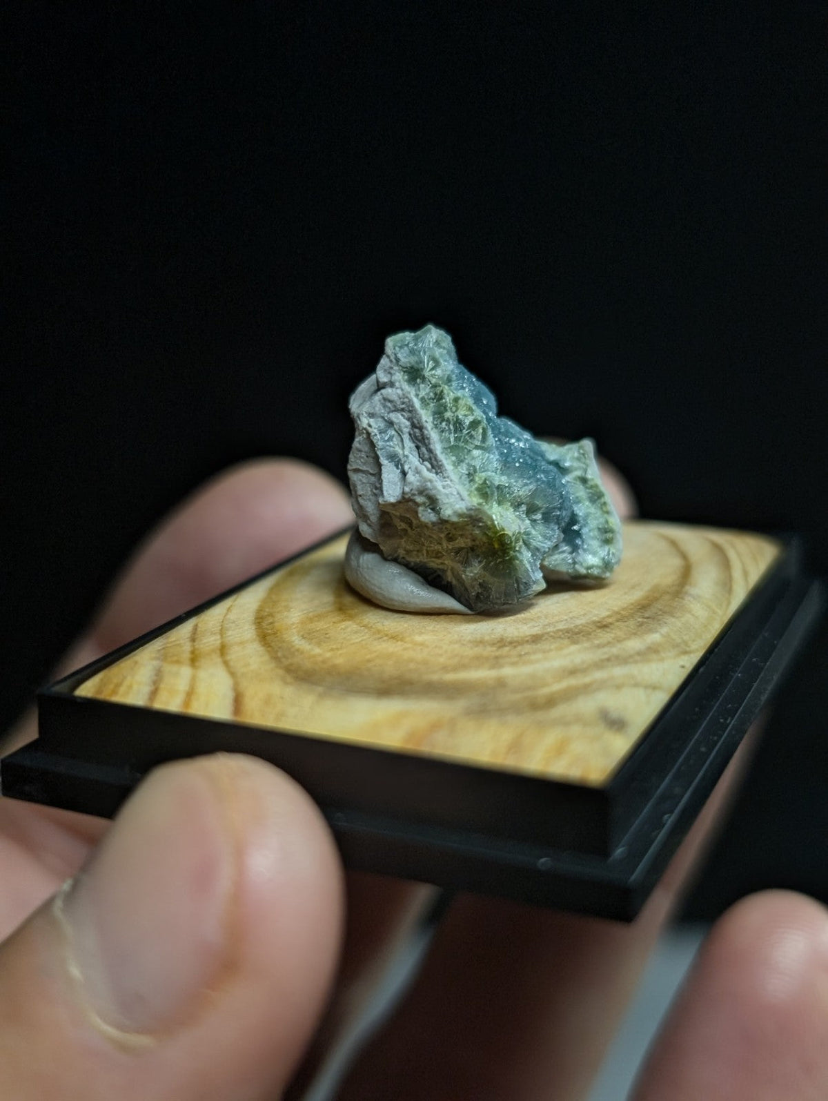 A small, irregularly shaped mineral specimen with a mix of green and gray hues rests on a wooden display base.