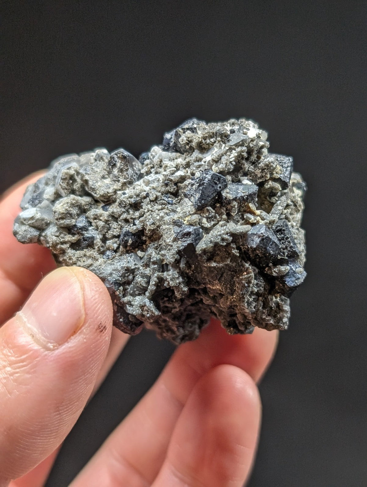 A hand holds a rough, irregularly shaped mineral specimen featuring dark gray to black crystalline formations embedded in a lighter gray matrix.