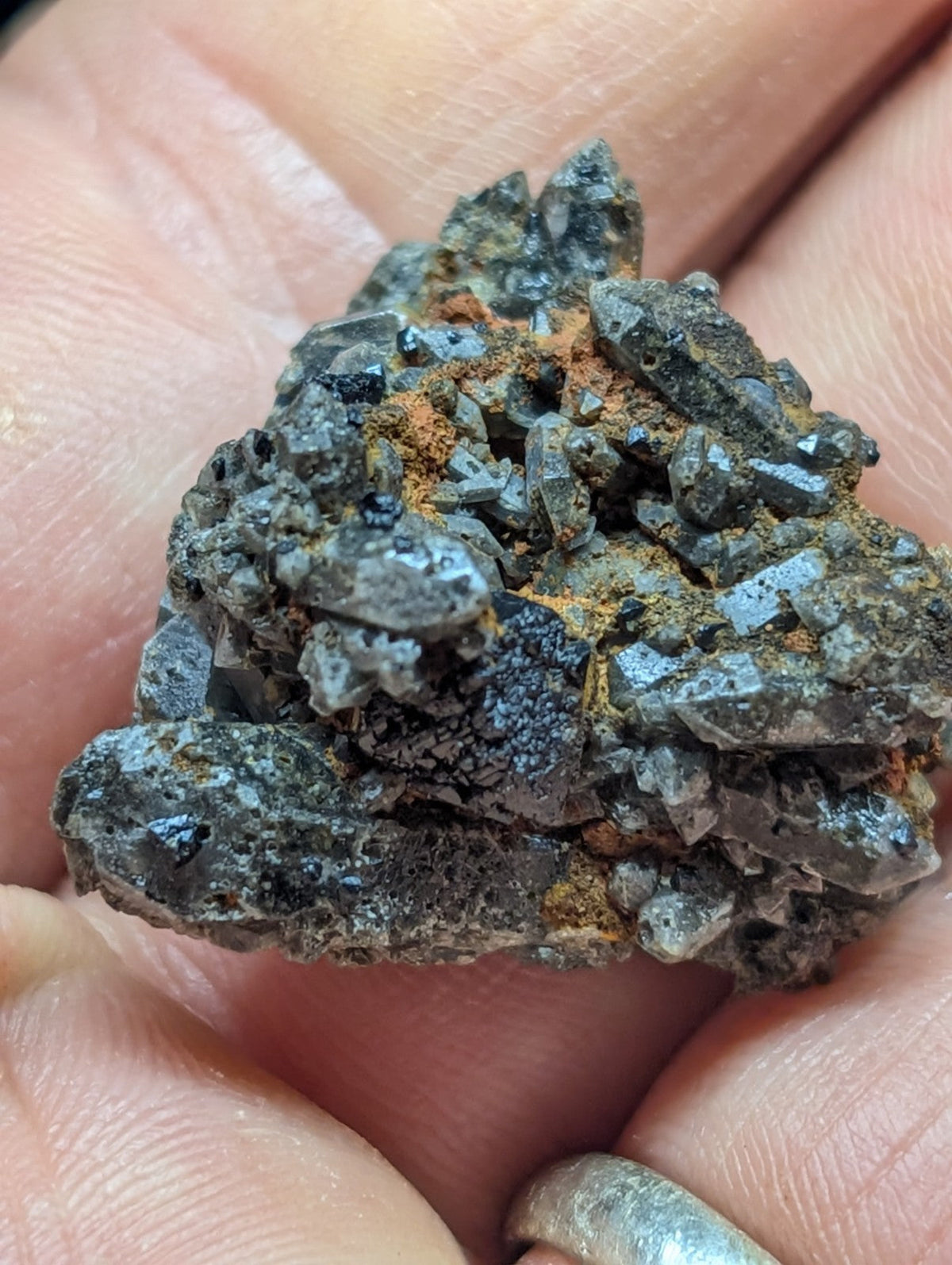 A hand holds a dark gray, crystalline mineral specimen with sharp facets and patches of orange rust-colored oxidation.