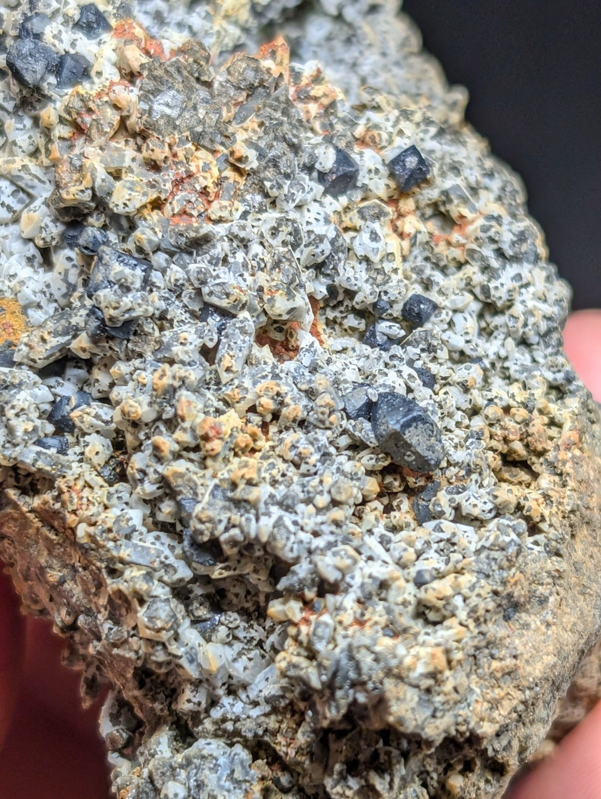 A rugged, textured mineral specimen featuring a mosaic of dark gray crystals embedded in a matrix of light gray and orange-brown earthy material.