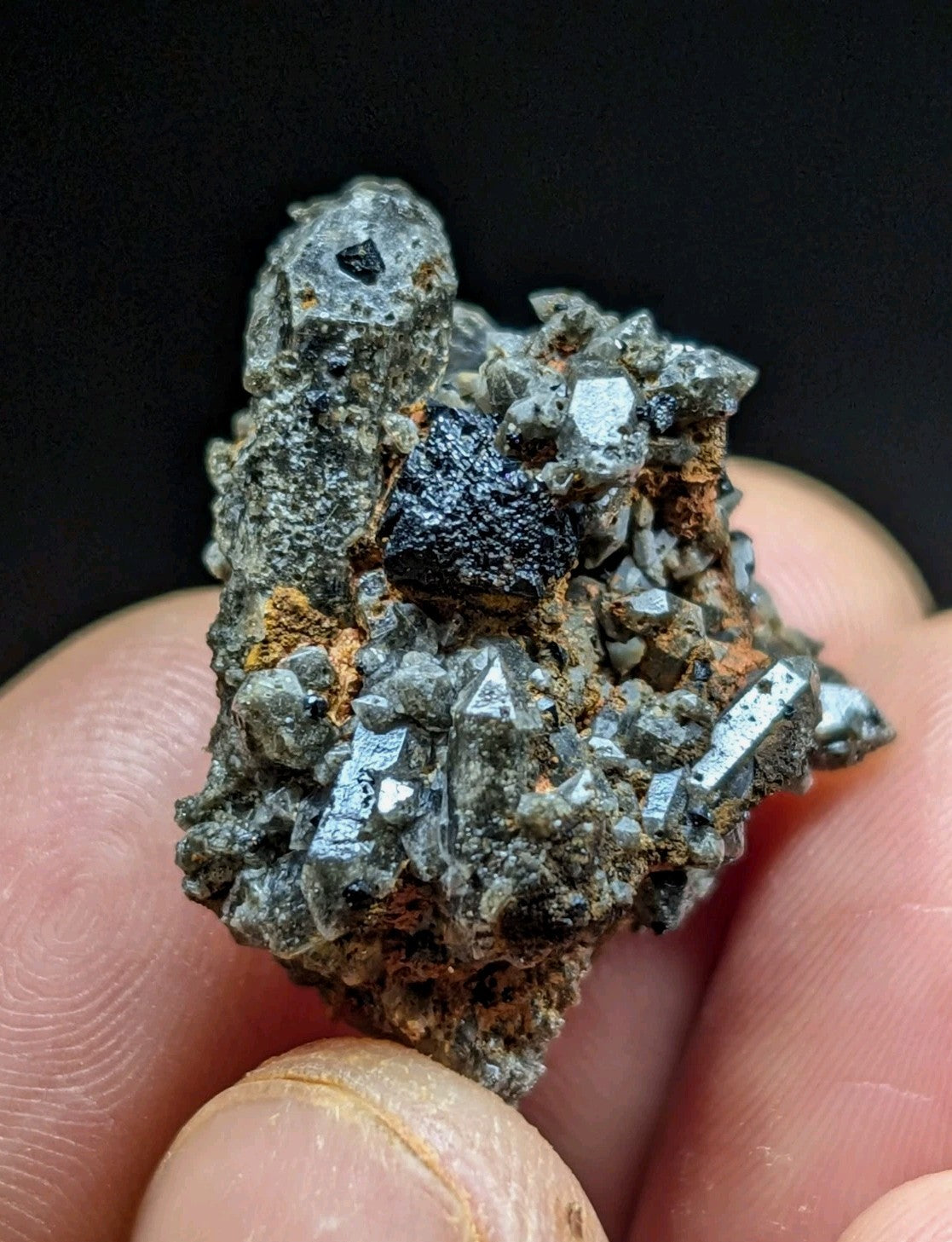 A hand holds a rough, irregularly shaped mineral specimen featuring prominent metallic gray crystals and a central dark, glossy black inclusion.