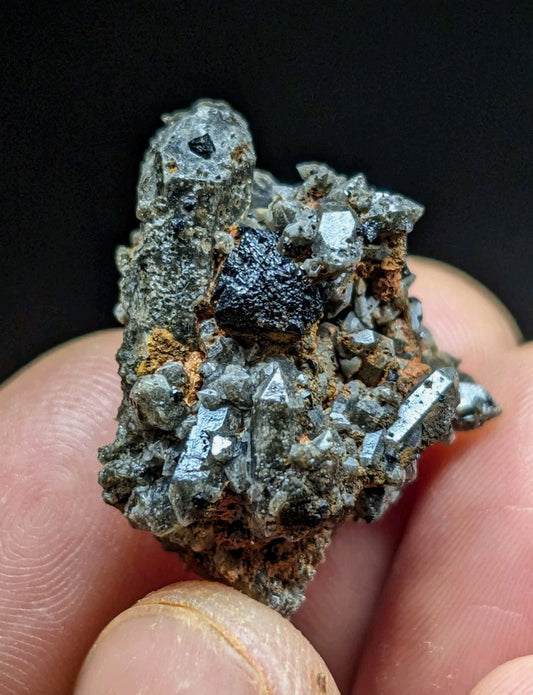 A hand holds a rough, irregularly shaped mineral specimen featuring prominent metallic gray crystals and a central dark, glossy black inclusion.