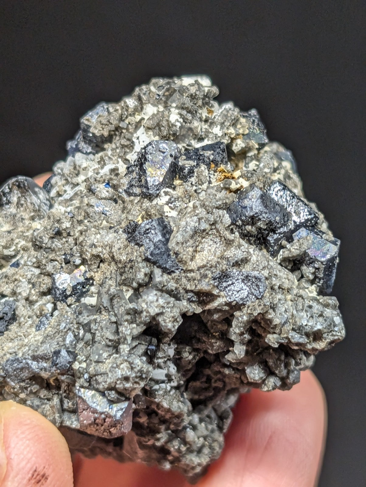 A hand holds a rough, irregularly shaped mineral specimen featuring dark metallic crystals embedded in a grayish matrix.