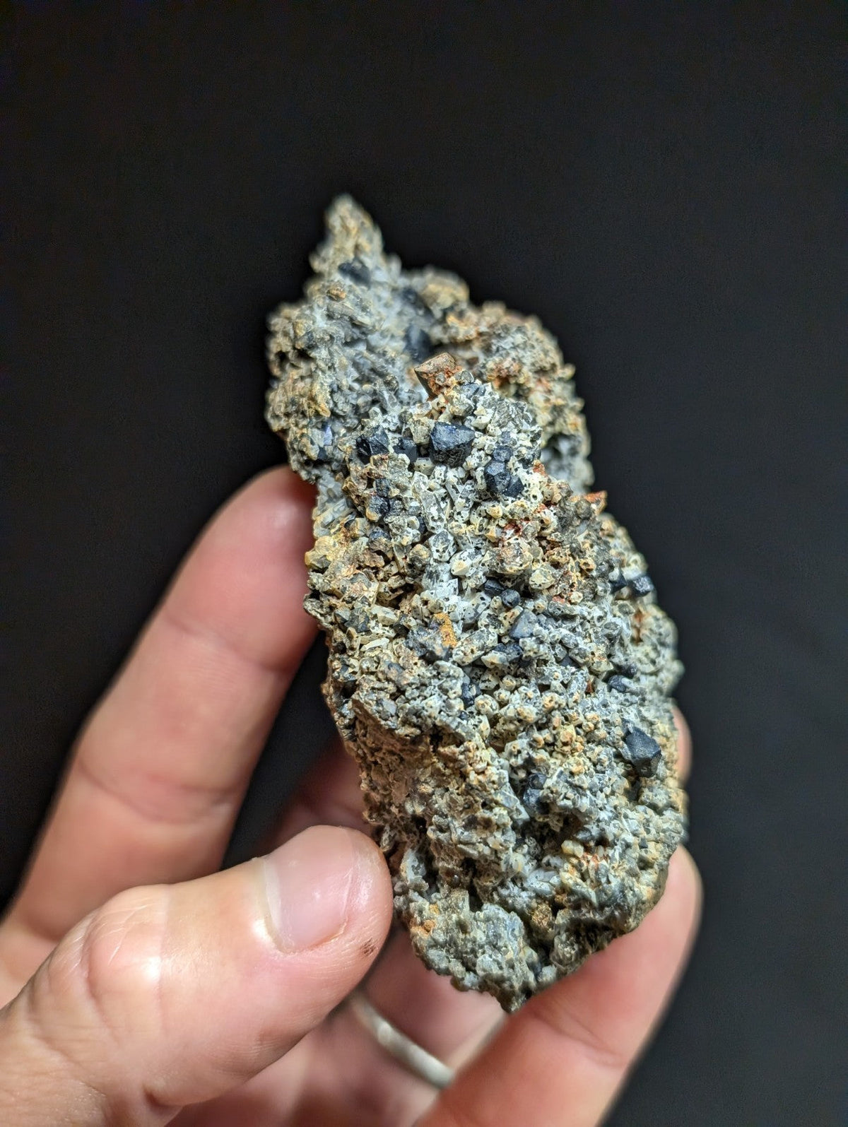A rough, irregularly shaped mineral specimen with a mottled gray and greenish surface, speckled with darker inclusions.