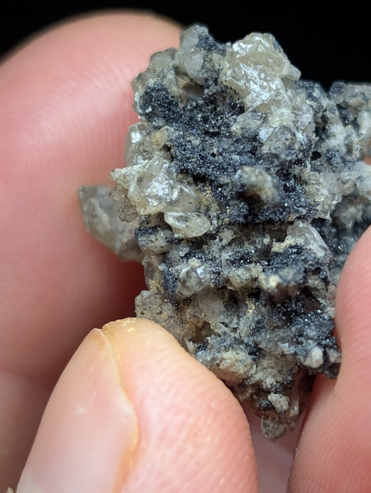 A hand holds a rough, irregularly shaped mineral specimen featuring translucent gray crystals intermingled with dark black speckles.