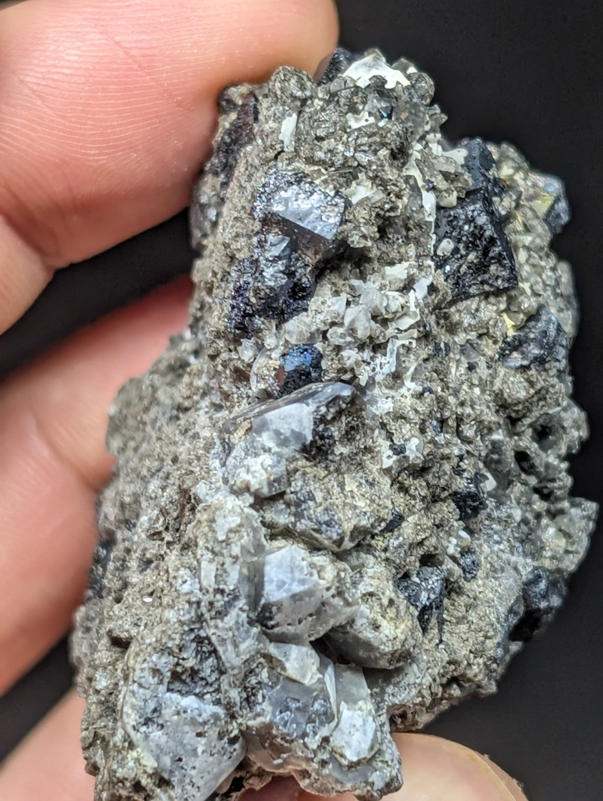 A hand holds a rough, grayish mineral specimen featuring prominent dark blue and translucent crystal formations embedded in a rocky matrix.