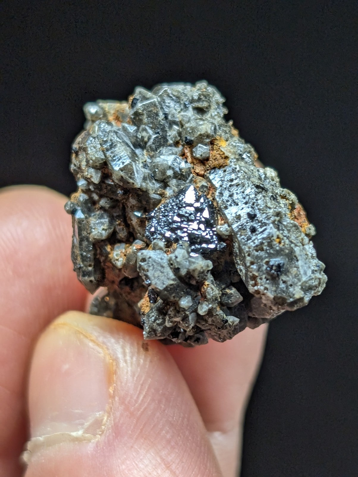 A cluster of metallic, crystalline minerals with a dark gray and black sheen, featuring sharp facets and embedded orange-brown oxidation.