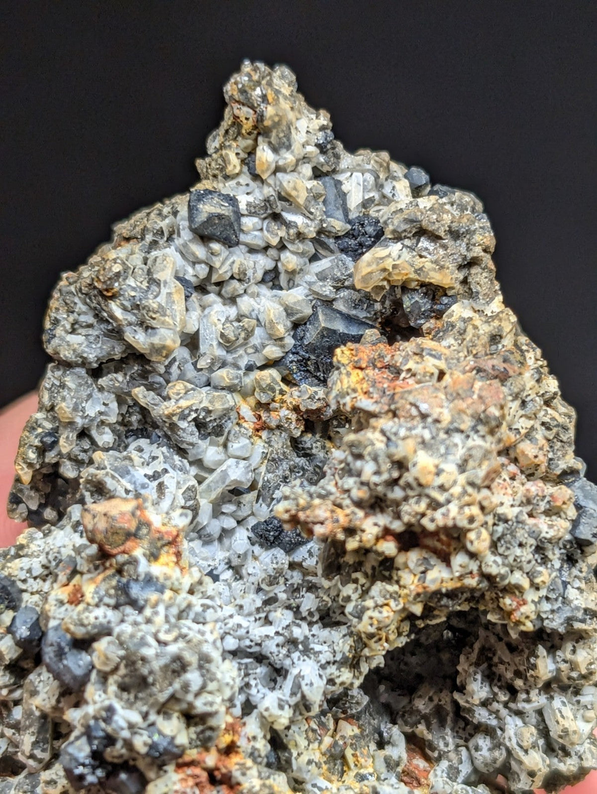 A rugged, multi-colored mineral specimen featuring a mix of gray and black crystalline formations with patches of orange rust-like material.