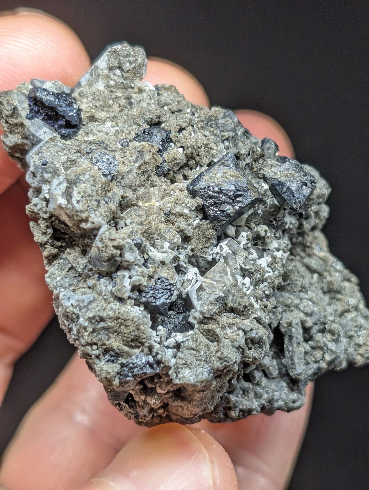 A hand holds a rough, grayish mineral specimen featuring dark, crystalline inclusions.
