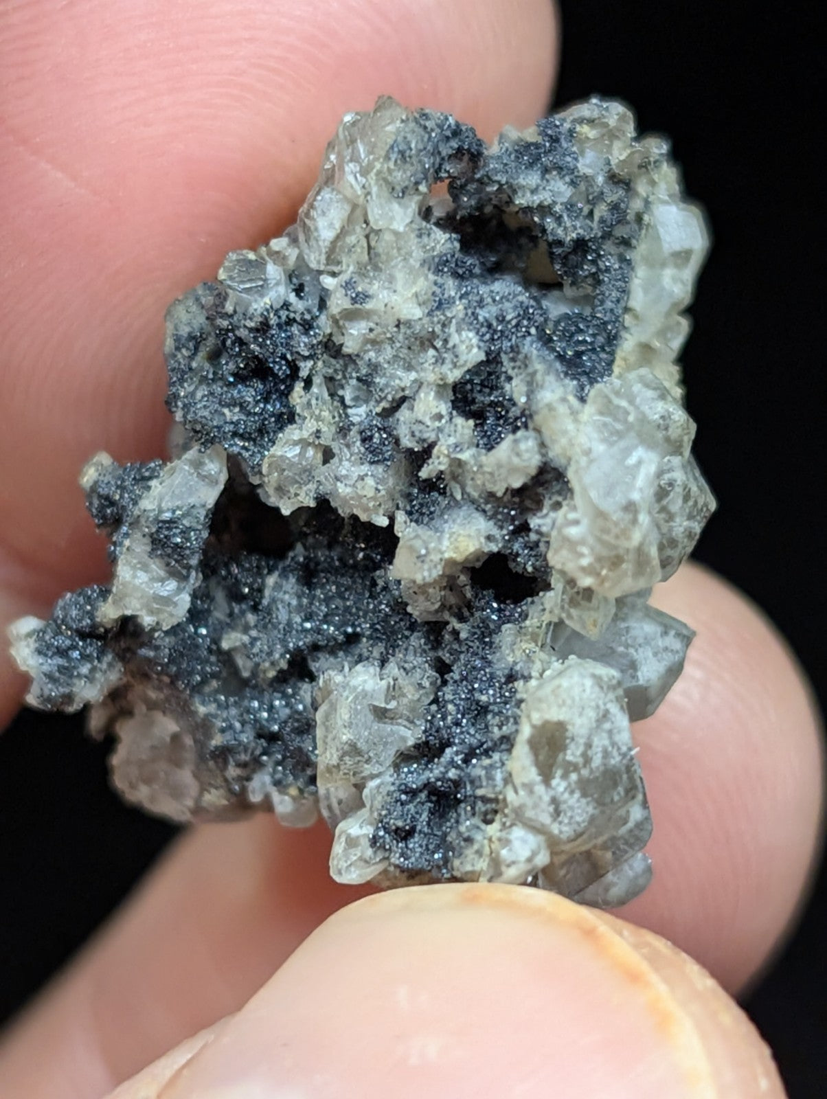 A hand holds a rough, irregularly shaped mineral specimen featuring translucent gray crystals intermingled with dark black speckles.