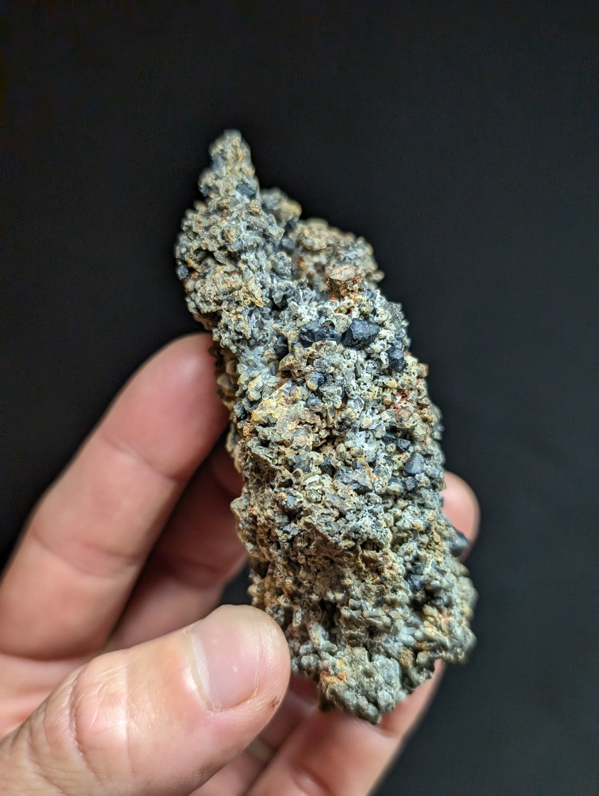 A rough, irregularly shaped mineral specimen with a mottled gray and greenish surface, featuring small dark inclusions.