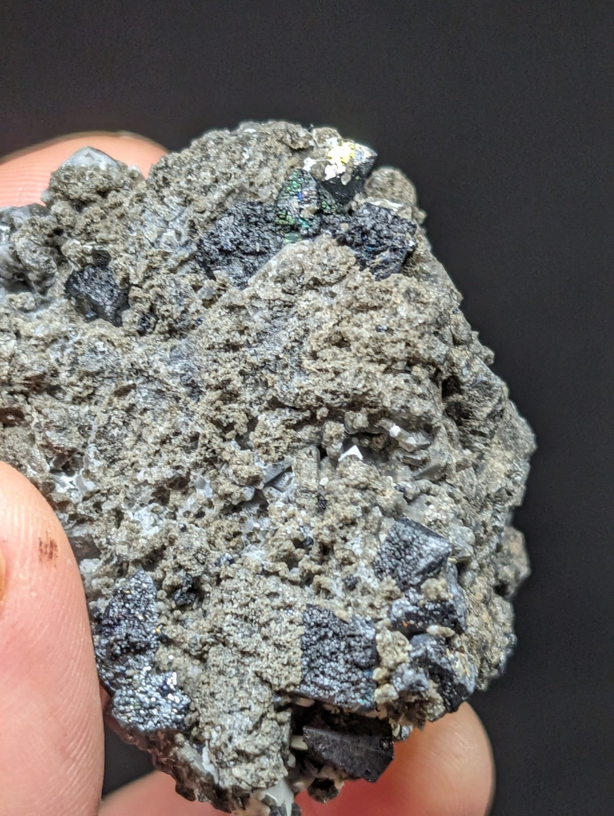 A rough, grayish mineral specimen with dark, crystalline inclusions and iridescent green-blue flecks.