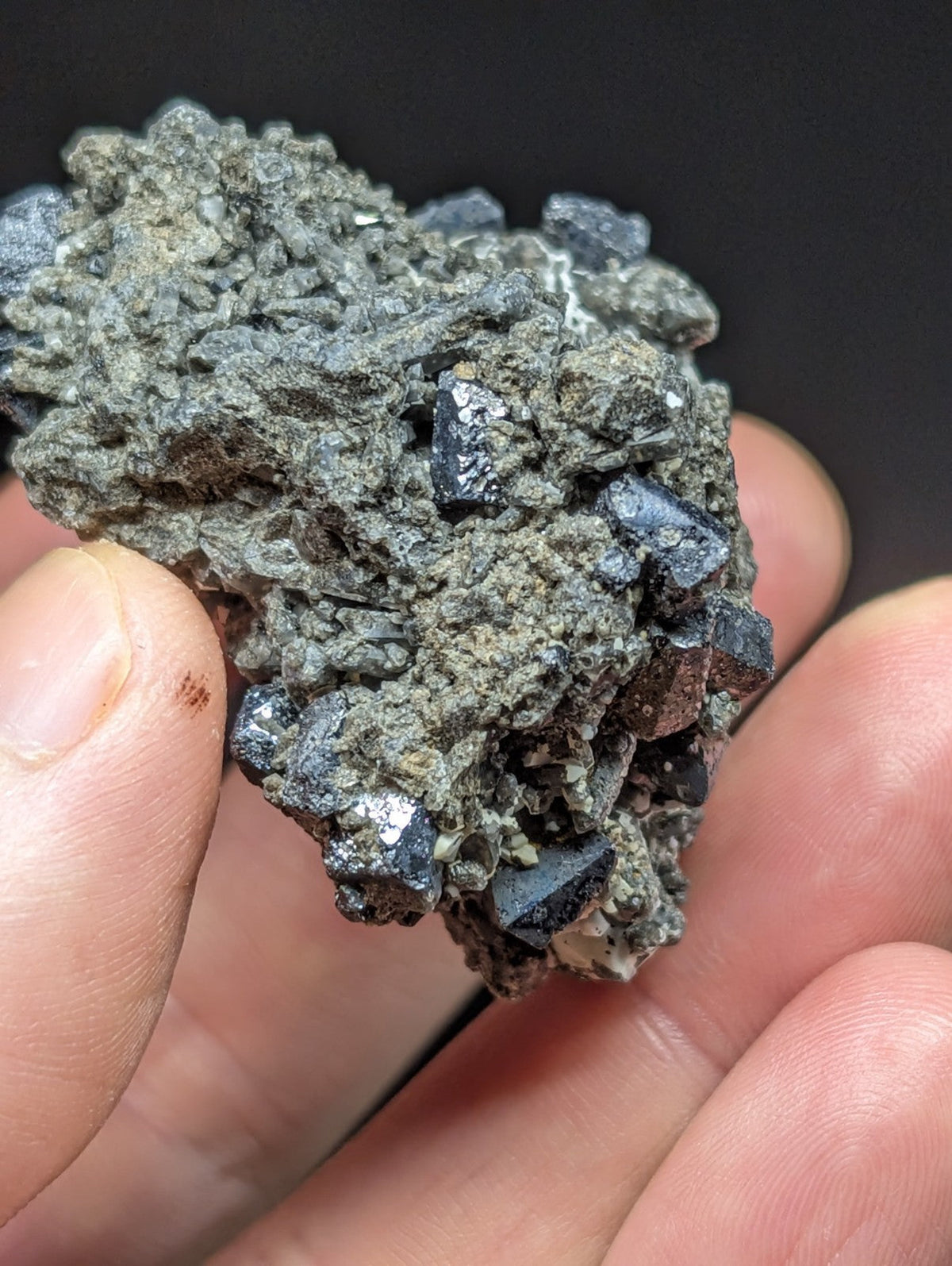 A hand holds a rough, grayish mineral specimen with dark blue to black crystalline formations embedded in its rocky matrix.