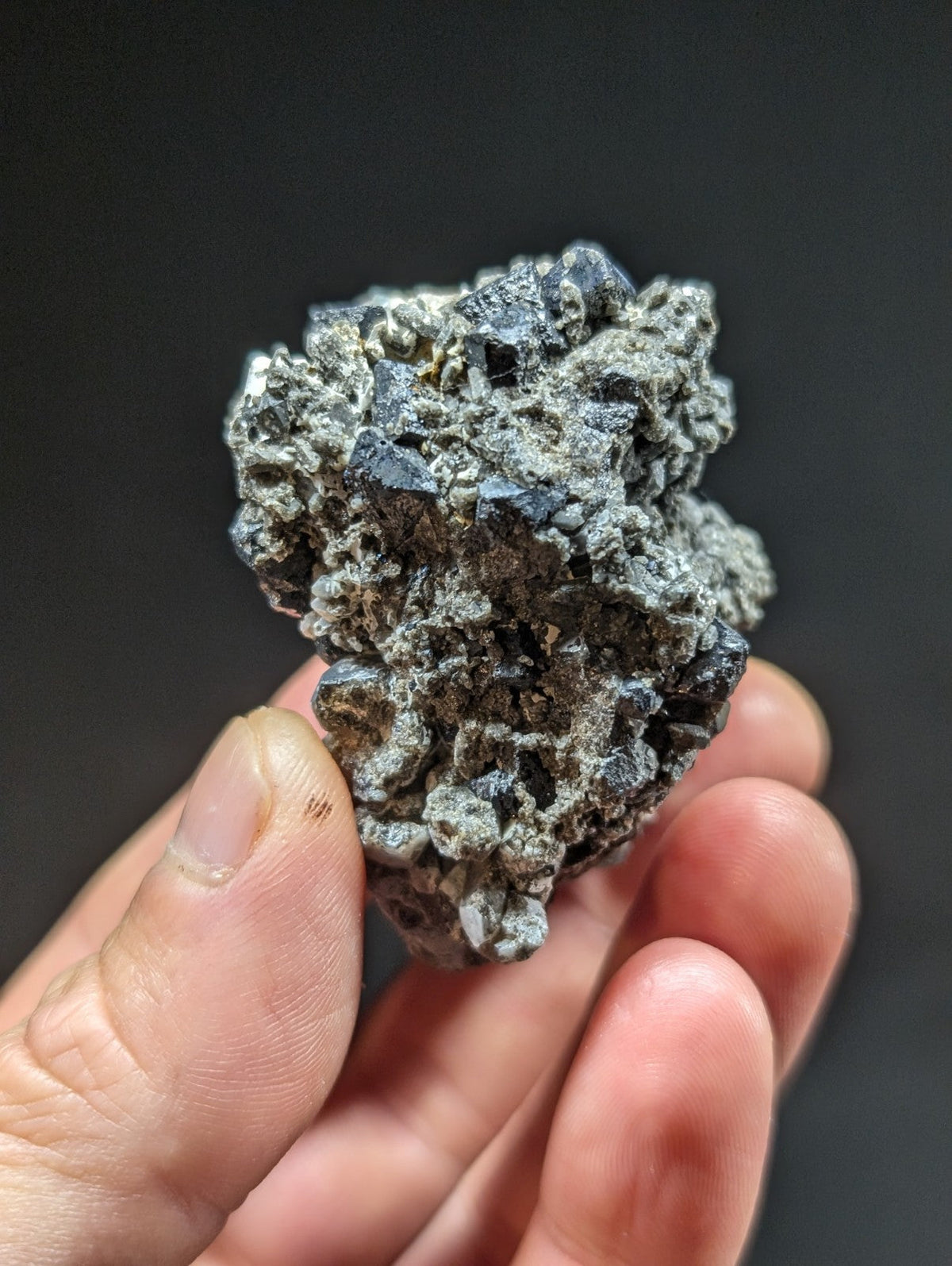 A rough, grayish-black mineral specimen with a clustered, crystalline texture held in someone’s hand.