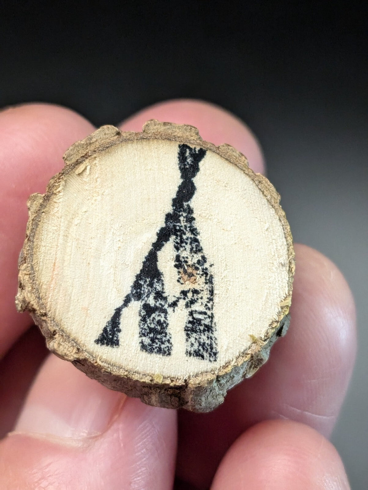 A hand holding a circular wooden slice with a black inked silhouette of an arrowhead design on its light-colored, textured surface.