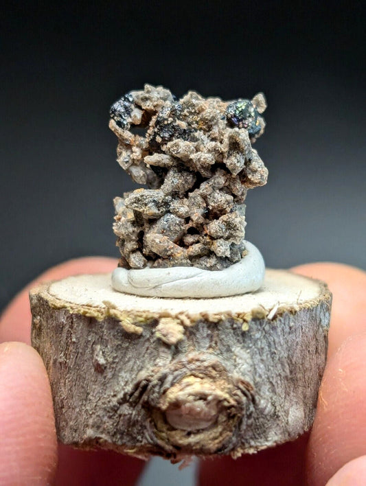 A rugged, earthy-toned mineral specimen with a rough, clustered texture and dark speckles sits atop a smooth white circular base mounted on a rustic wooden slice.
