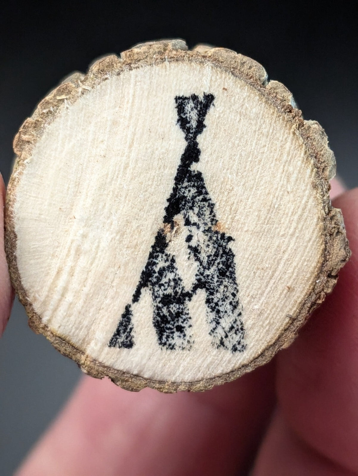 A circular wooden slice with a natural light beige surface and rough, brown bark edge, featuring a black charcoal-burned silhouette of an a-frame tent.