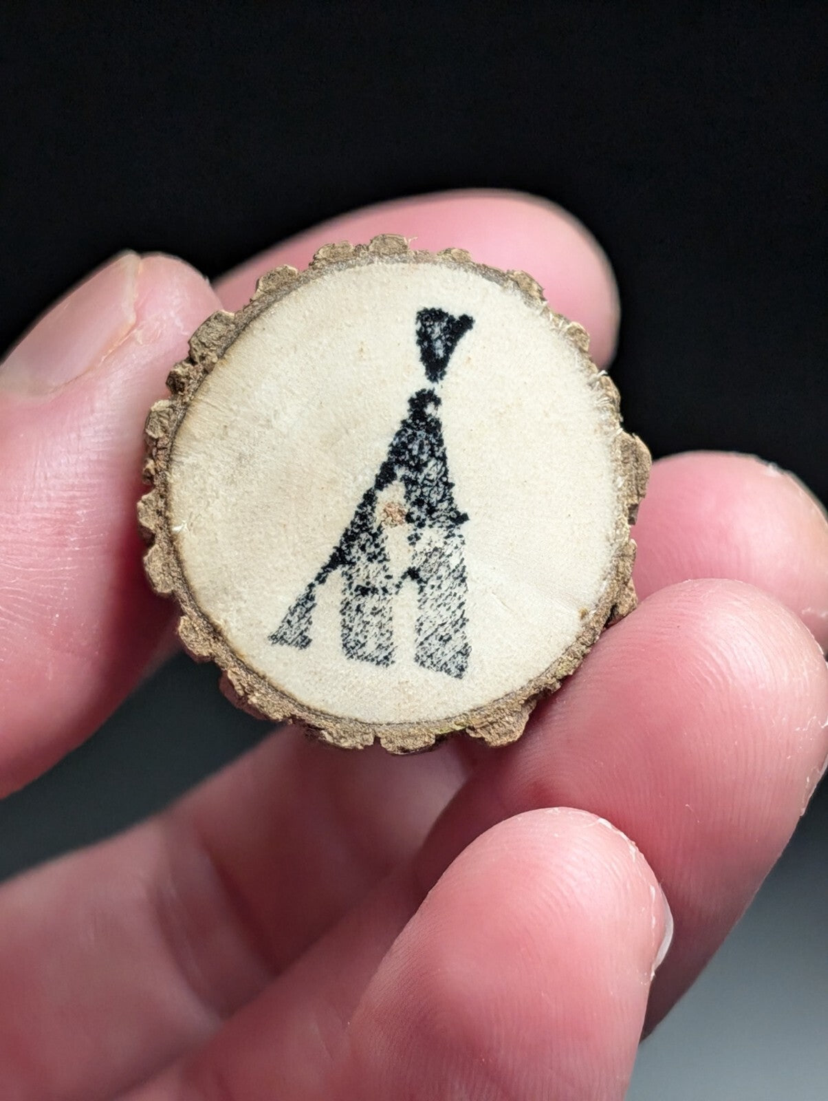 A circular wooden slice pendant featuring a black embroidered teepee design on its light beige surface, framed by the natural, textured bark edge.