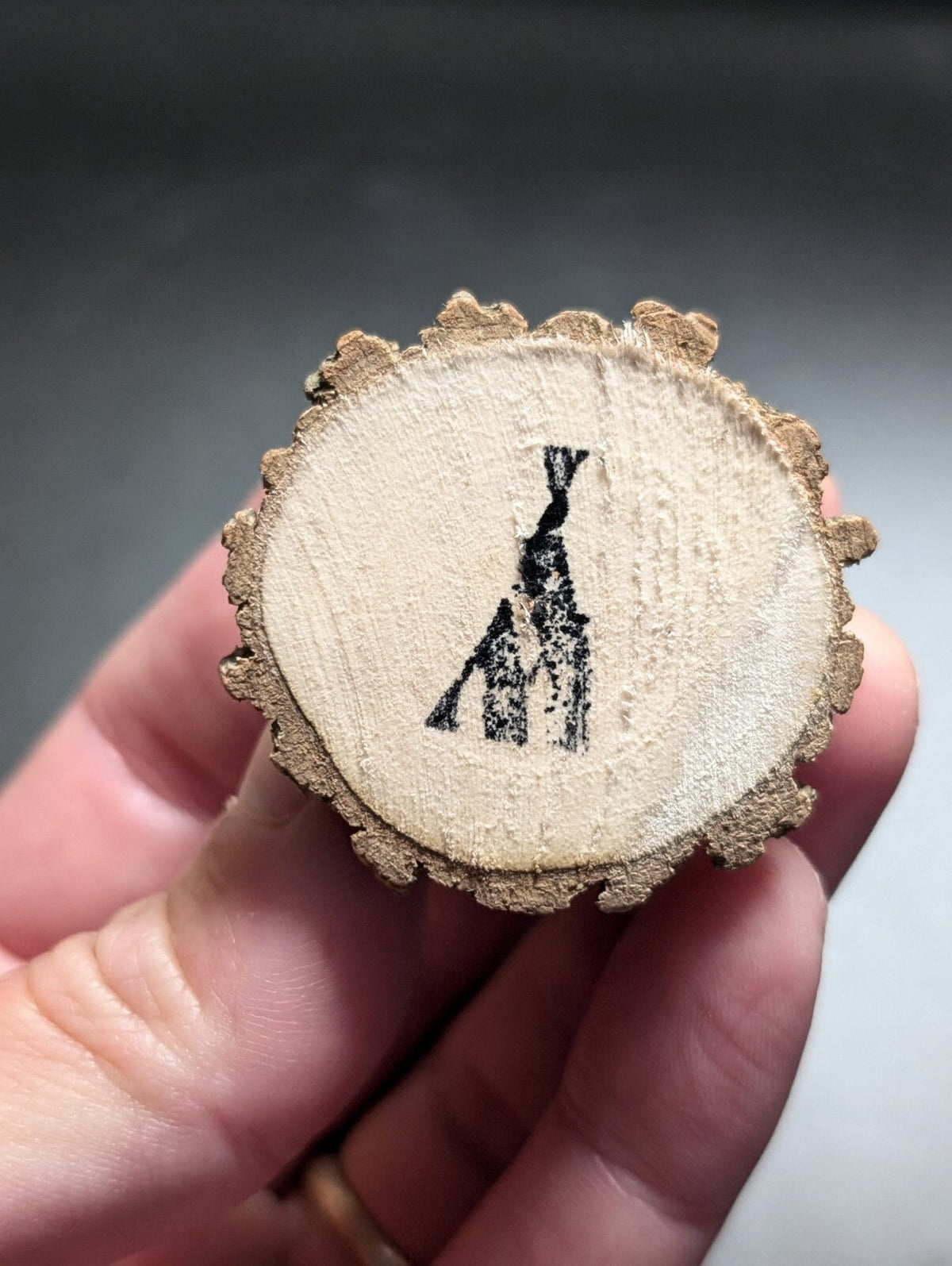 A circular, light brown wooden slice with a black inked silhouette of a teepee design on its flat surface.