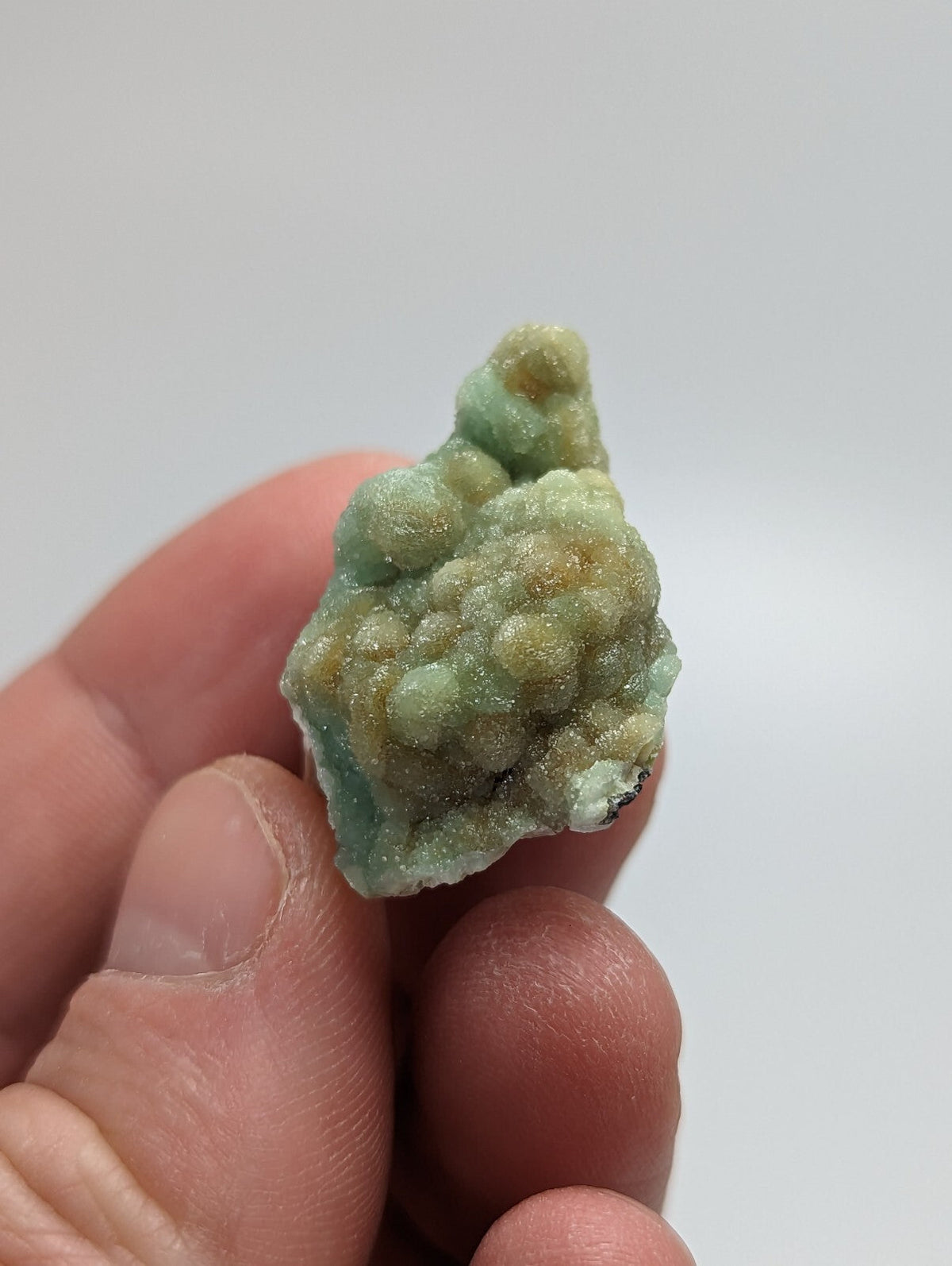 A hand holds a small, irregularly shaped mineral specimen with a textured surface featuring clusters of light green and yellowish-brown hues.