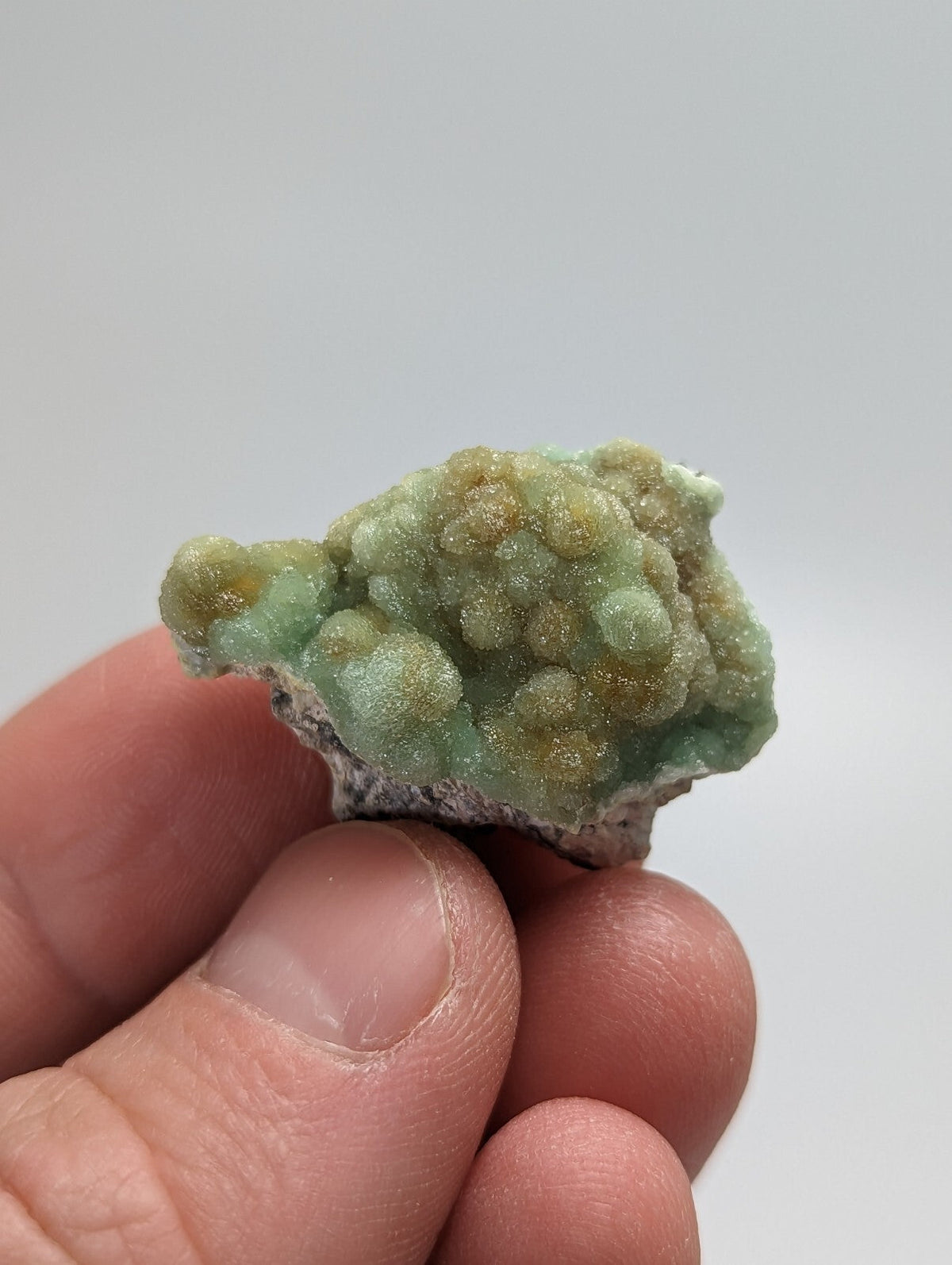 A hand holds a small, irregularly shaped mineral specimen featuring clusters of translucent green crystals with golden-yellow speckles and a rough, textured surface.