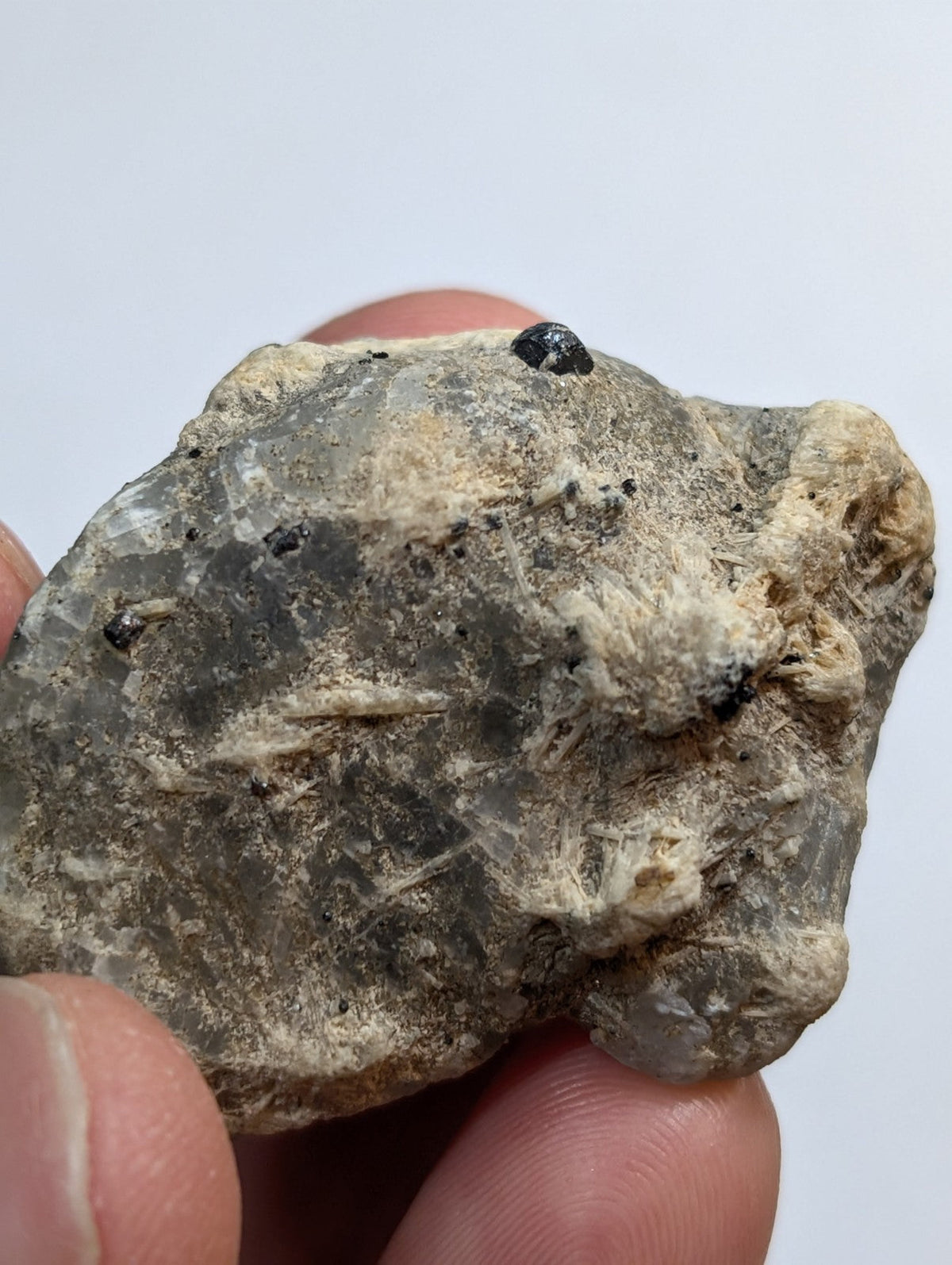 A rough, grayish-green mineral specimen with visible crystalline textures and small black inclusions.