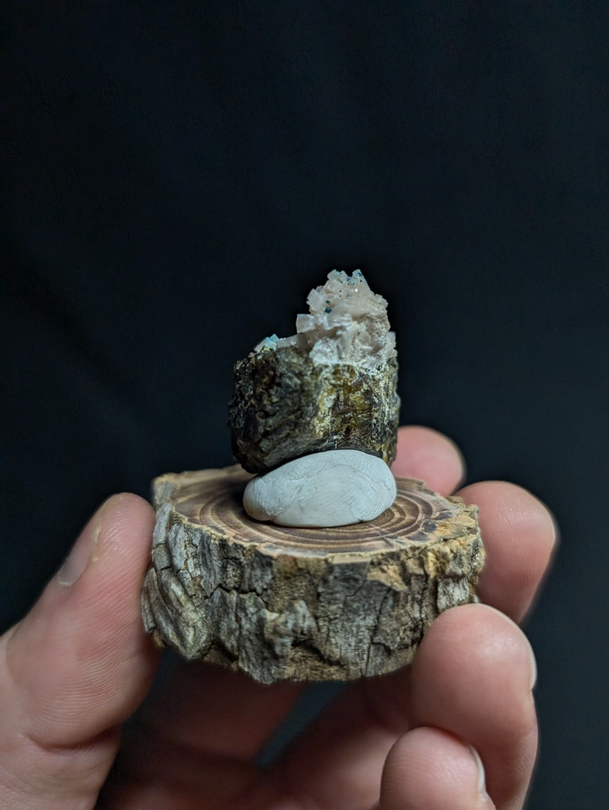 A hand holds a rustic wooden slice with natural bark edges, topped by a smooth white stone and a textured mineral specimen featuring greenish hues and crystalline formations.