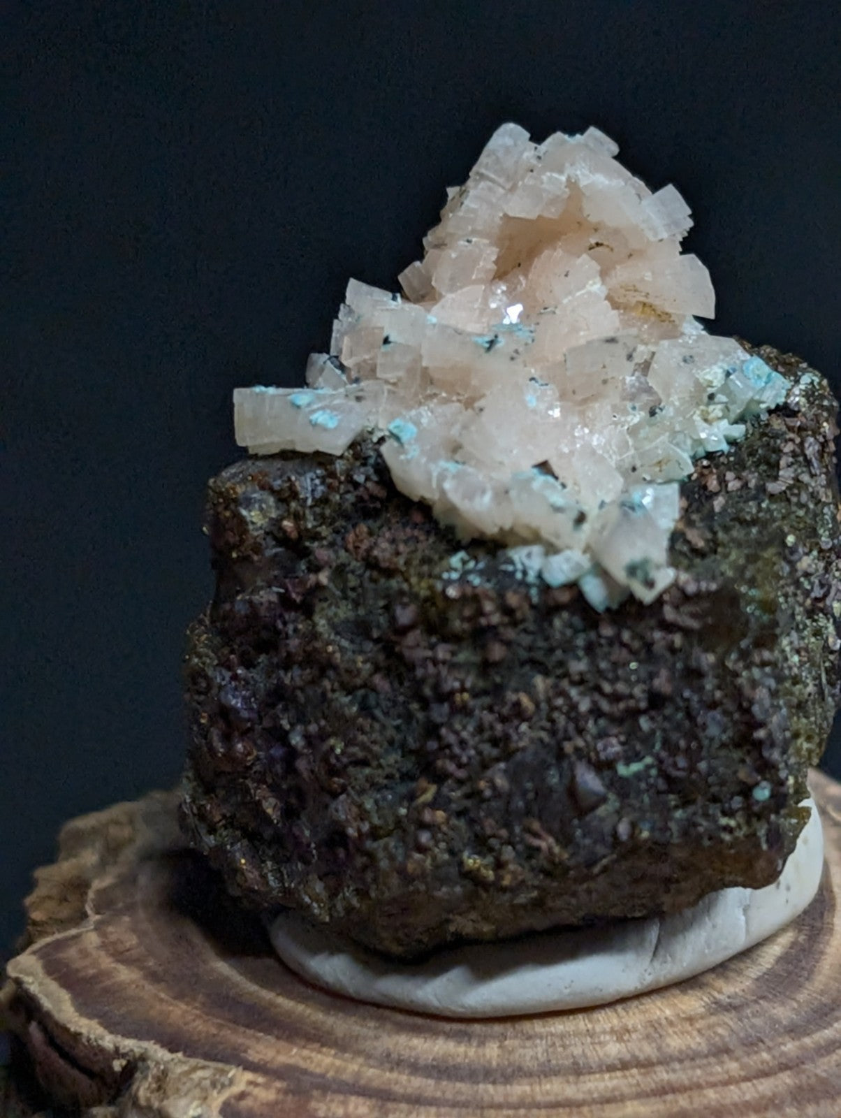 A translucent, crystalline mineral formation with a pale pinkish hue and scattered blue-green flecks sits atop a dark, rugged rock base.