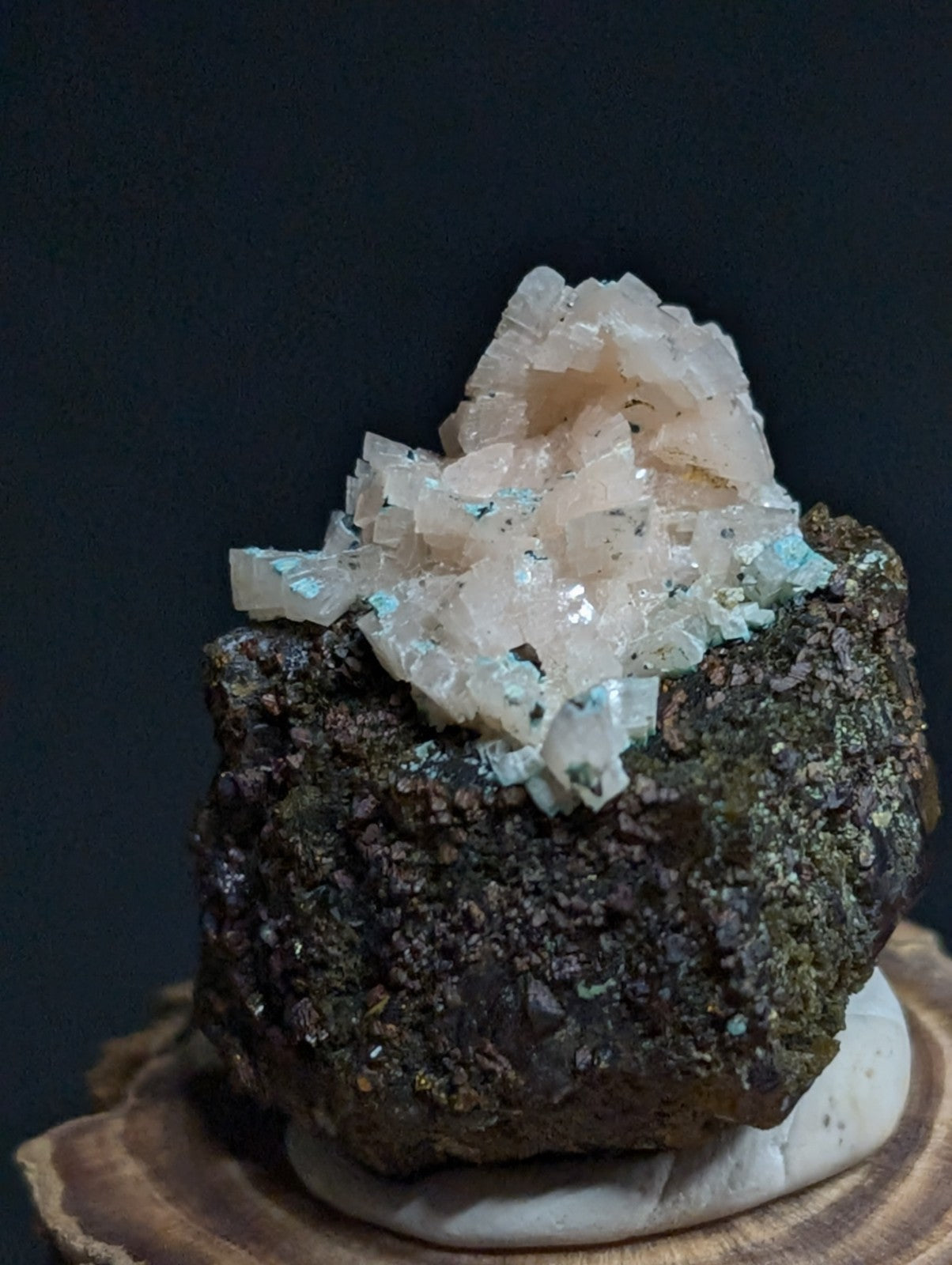 A translucent, crystalline mineral formation with a pale pinkish hue and subtle blue-green flecks sits atop a dark, rough-textured rock base.