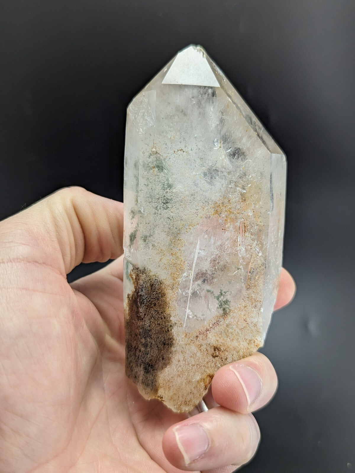 A clear quartz crystal with natural inclusions and earthy brownish streaks, held gently in a hand against a dark background.