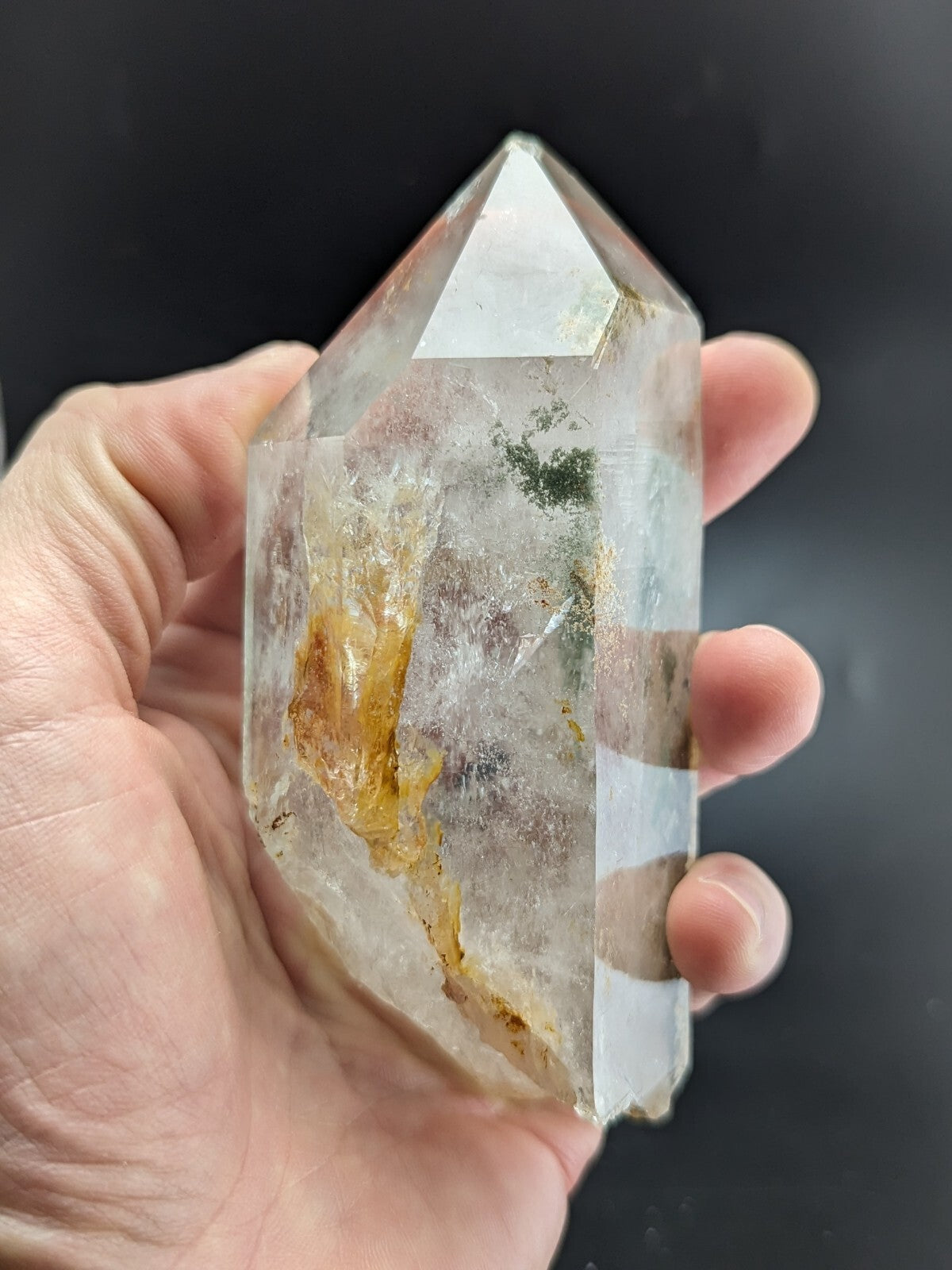 A clear quartz crystal with natural inclusions of golden and greenish hues, held gently in a hand.