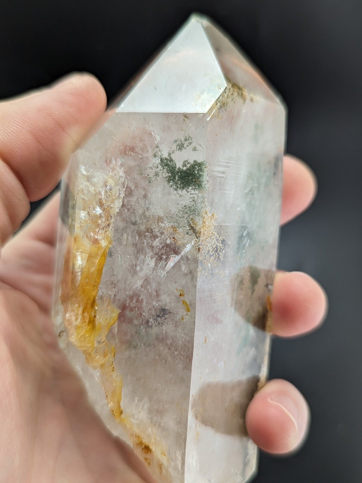 A translucent quartz crystal with natural inclusions of green and yellow mineral deposits, held gently in a hand.